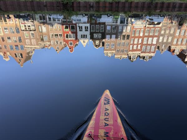 This serene photo captures the view from a paddleboard gliding along the Geldersekade in Amsterdam, with the iconic canal houses beautifully reflected in the still blue water